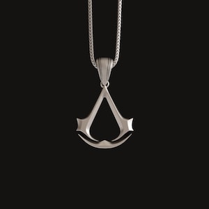Könnte beinhalten: Eine silberne Halskette mit einem Anhänger in Form des Assassin's Creed-Logos.