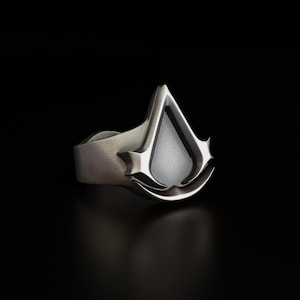 Könnte beinhalten: Ein silberner Ring mit einem stilisierten, dreidimensionalen Assassin's Creed-Logo. Das Logo ist eine stilisierte, spitze, tropfenförmige Form mit einem versteckten Klingen-Design.