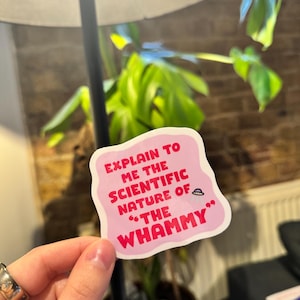 May include: A pink sticker with white text: "EXPLAIN TO ME THE SCIENTIFIC NATURE OF 'THE WHAMMY'". The sticker is held in front of a houseplant and a lamp.