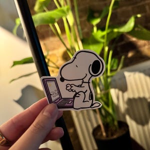Może przedstawiać: Biała naklejka Snoopy z czarnymi detalami, trzymająca laptopa z grafiką Snoopy na ekranie. Naklejka ma fioletową obwódkę i jest trzymana przed rośliną.