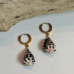 May include: A pair of gold-toned hoop earrings featuring dangling cone-shaped seashells. Each shell has a white base with dark brown, leopard-like spots and a small, centered white pearl accent. The hoops are polished gold metal.