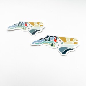 K&ouml;nnte beinhalten: Zwei Aufkleber in Form des Bundesstaates North Carolina mit einem farbenfrohen Design, das Berge, einen Kardinal, eine Sonne und die Worte "North Carolina" zeigt.