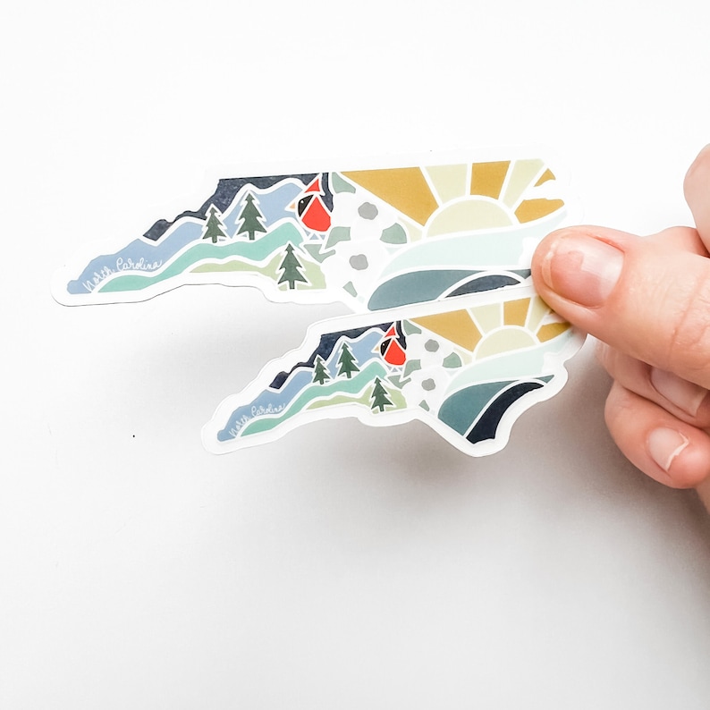 K&ouml;nnte beinhalten: Zwei Aufkleber in Form des Bundesstaates North Carolina mit einem farbenfrohen Design, das einen Sonnenaufgang, Berge, B&auml;ume und einen Kardinal zeigt. Die Aufkleber zeigen den Schriftzug "Visit Carolina" in einer handschriftlichen Schrift.