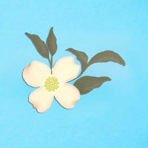 May include: A flat, die-cut illustration of a dogwood flower with four pale cream petals featuring soft pink-tinted edges and a central cluster of light green dots, accompanied by dark olive green leaves on a bright blue background.