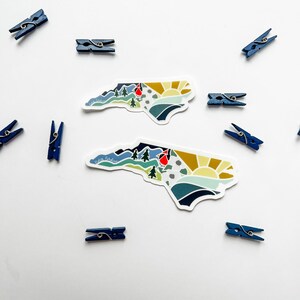 K&ouml;nnte beinhalten: Zwei bunte Aufkleber mit der Umrissform des Bundesstaates North Carolina, die ein Berg-, Sonnen- und Kardinalvogel-Design zeigen. Die Aufkleber befinden sich auf einem wei&szlig;en Hintergrund mit blauen W&auml;scheklammern.