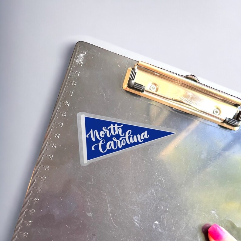 May include: A blue pennant with white text that reads "North Carolina" on a silver clipboard.
