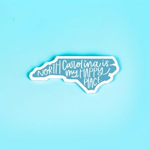 May include: A light blue sticker in the shape of North Carolina with the text "North Carolina is my HAPPY PLACE" in white script. The sticker has a white border and is set against a light blue background.
