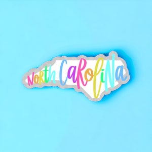 May include: A white outline of the state of North Carolina with the words "North Carolina" written in a rainbow of colors.
