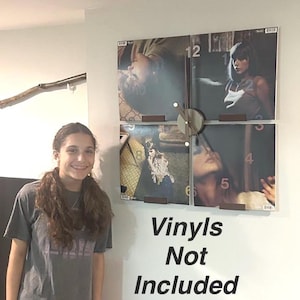 May include: A clock made from four vinyl records with a wooden frame. The clock is mounted on a white wall. The clock is surrounded by four vinyl records. The text "vinyls not included" is written below the clock.