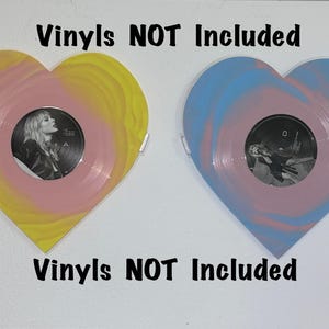 May include: Two heart-shaped vinyl record display stands. The stands are painted in a pink and yellow swirl pattern and a pink and blue swirl pattern. The text "Vinyl NOT Included" is printed on the white background.