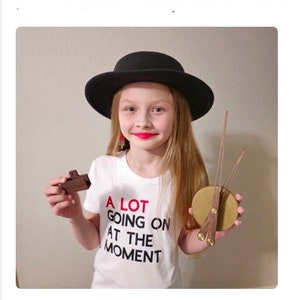 May include: A young person wearing a white t-shirt with the text "A LOT GOING ON AT THE MOMENT" in black and red letters. They are wearing a black bowler hat and holding a small brown object in one hand and a gold and brown clock in the other.