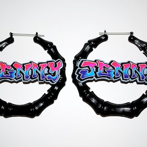 May include: Pair of black bamboo-style hoop earrings. Each earring features the name "JENNY" in a colorful graffiti-style font with pink, blue, and purple hues. The earrings are set against a white background.
