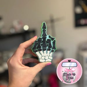 May include: A decorative skeleton hand with the middle finger raised, covered in glitter. The hand is black and white with green and blue highlights. Likely a car accessory or ornament.
