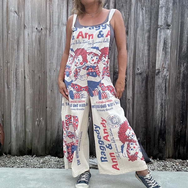 May include: Cream-colored Raggedy Ann & Andy jumpsuit with red, blue, and white graphics. The jumpsuit features the text "Raggedy Ann & Andy" and "The original doll with her" on the front. The straps are white, and the pants are wide-legged.