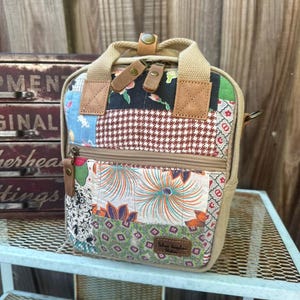 May include: A small, patchwork fabric backpack with a tan canvas body and straps. The patchwork includes floral, geometric, and patterned fabrics in various colors. The bag has a zipper closure and leather accents.
