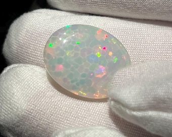 May include: A white opal cabochon with a speckled pattern of iridescent green, pink, and blue colors.