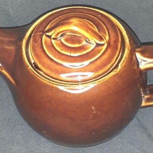 May include: A brown ceramic teapot with a lid featuring concentric circles. The teapot has a spout, a handle, and a rounded body. The lid has a decorative design. The teapot is a warm, earthy brown color.