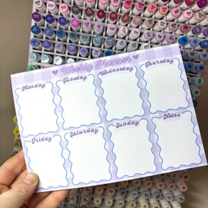 May include: A weekly planner with a light purple gingham border and wavy blue edges. The planner has sections for each day of the week, plus a notes section. The text "Weekly Planner" is at the top, with the days of the week labeled below.