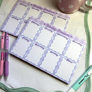 May include: Two weekly planner notepads with a light purple wavy border and a gingham pattern. Each notepad has sections for each day of the week and notes. Pens and a highlighter are also visible.
