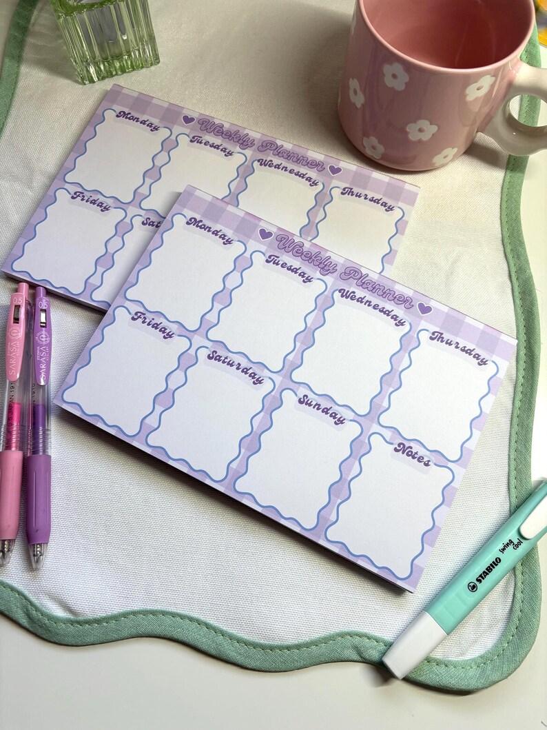 May include: Two weekly planner notepads with a light purple wavy border and a gingham pattern. Each notepad has sections for each day of the week and notes. Pens and a highlighter are also visible.