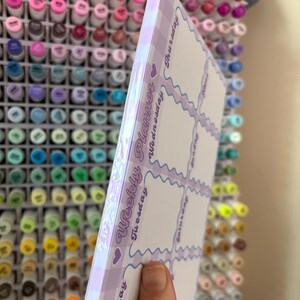 May include: A weekly planner with a lilac gingham border and a white interior. The planner has a wavy design and the days of the week are printed vertically on the side. The words "Weekly Planner" are printed on the side.