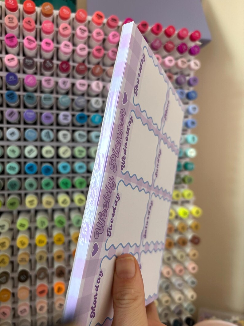 May include: A weekly planner with a lilac gingham border and a white interior. The planner has a wavy design and the days of the week are printed vertically on the side. The words "Weekly Planner" are printed on the side.
