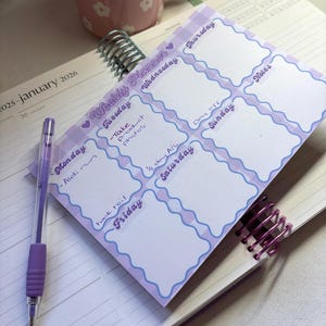 May include: A weekly planner with a purple and white gingham border, featuring days of the week and space for notes. A purple pen rests on the planner, and a pink floral mug is in the background. The planner is open to January 2026.