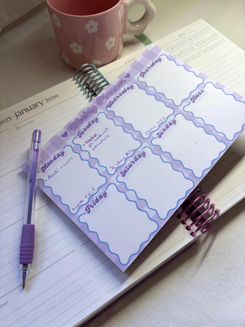 May include: A weekly planner with a purple and white gingham border, featuring days of the week and space for notes. A purple pen rests on the planner, and a pink floral mug is in the background. The planner is open to January 2026.