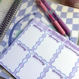 May include: A weekly planner with a purple and white checkered design, featuring a wavy border and labeled days of the week. Two pens, one pink and one purple, rest on top. A spiral-bound notebook and a floral mug are in the background.