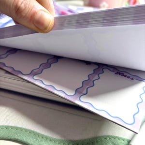 May include: A hand flips through the pages of a notepad. The notepad has a white background with light purple and blue wavy borders. The word "Notes" is visible on one page. The notepad rests on a green fabric.