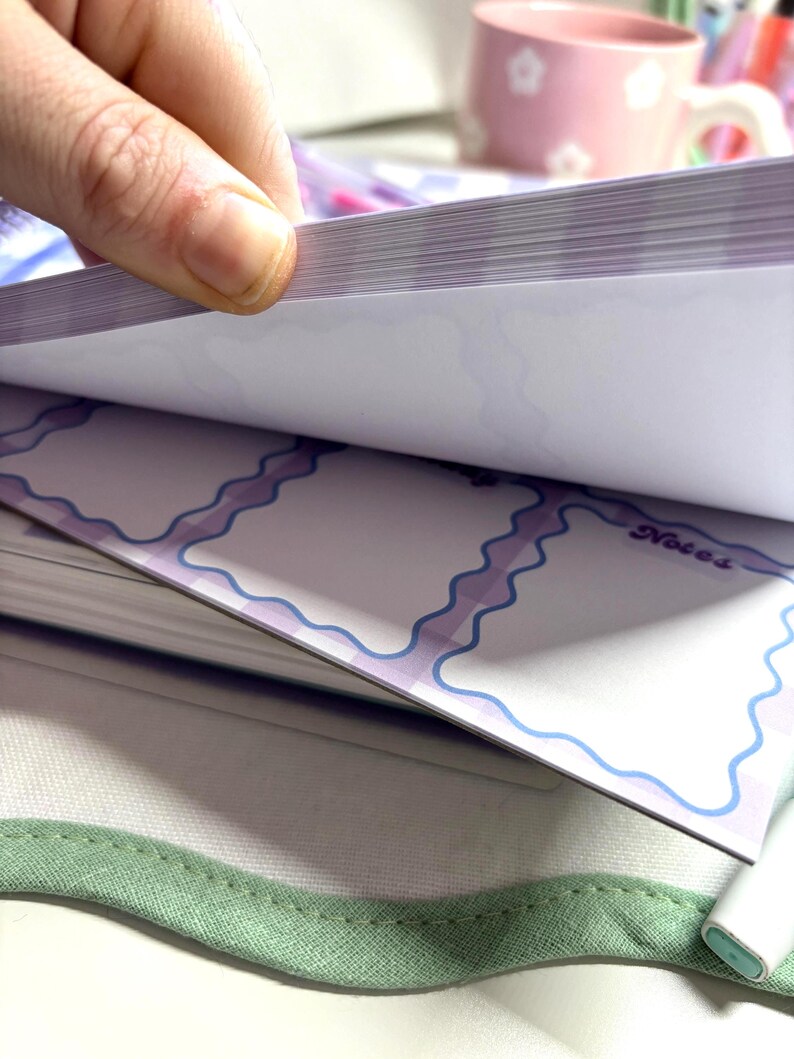 May include: A hand flips through the pages of a notepad. The notepad has a white background with light purple and blue wavy borders. The word "Notes" is visible on one page. The notepad rests on a green fabric.