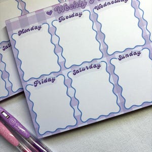 May include: A stack of weekly planner notepads with a purple gingham pattern and wavy blue borders. Each notepad features spaces for each day of the week, with the days labeled in a cursive font. Two pens, one pink and one purple, are in the foreground.