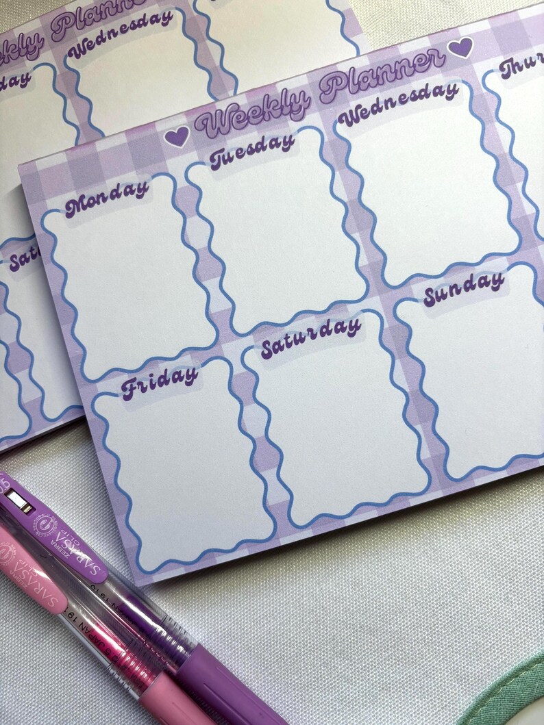 May include: A stack of weekly planner notepads with a purple gingham pattern and wavy blue borders. Each notepad features spaces for each day of the week, with the days labeled in a cursive font. Two pens, one pink and one purple, are in the foreground.