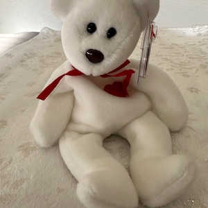 Könnte beinhalten: Weißer Plüschteddybär mit einem roten Band um den Hals und einem roten Herzen auf der Brust. Der Bär hat schwarze Augen und eine braune Nase. Der Bär sitzt auf einer weißen, flauschigen Oberfläche.