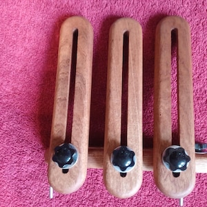 May include: Three wooden tools with a dark finish, each featuring a long, curved slot and a black knob. The tools are connected by a horizontal wooden piece, and sit on a pink surface. The tools have a metal pin at the bottom.