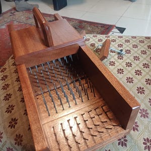 May include: A wooden flax hackle with a handle and metal tines. The hackle is made of dark brown wood and is designed for processing flax fibers. The tines are evenly spaced and extend upwards from the base.