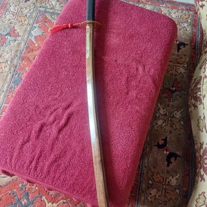 May include: A wooden katana sword with a black handle and a red tassel. The sword is lying on a red towel.