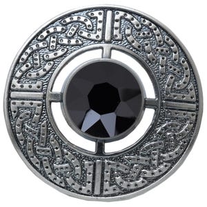 Puede incluir: Un broche redondo plateado con una gema negra central. El broche presenta un intrincado dise&ntilde;o de nudo celta alrededor de la gema y el borde exterior. El dise&ntilde;o es detallado y sim&eacute;trico, con un acabado pulido.