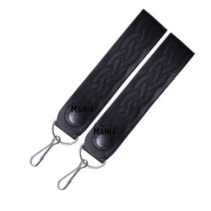 May include: Two black leather kilt straps with a Celtic knot design and metal clips. The straps are labeled "Kilt Mania".