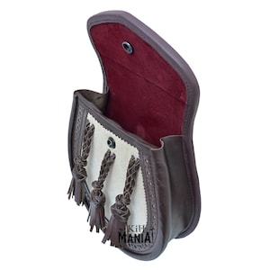 May include: A brown leather bag with a white leather flap and three braided leather tassels. The bag is open and the inside is lined with red fabric.
