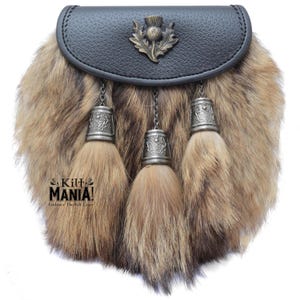 Puede incluir: Un sporran tradicional con una parte superior de cuero negro y un cuerpo de piel. El sporran presenta tres borlas decorativas con tapas y cadenas de metal plateado. Un emblema de cardo adorna la parte delantera. El texto "Kilt MANIA! Embrace The Kilt Craze" es visible.