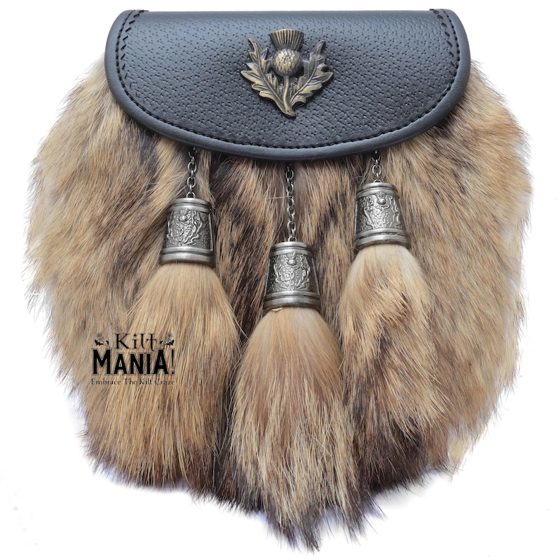Puede incluir: Un sporran tradicional con una parte superior de cuero negro y un cuerpo de piel. El sporran presenta tres borlas decorativas con tapas y cadenas de metal plateado. Un emblema de cardo adorna la parte delantera. El texto "Kilt MANIA! Embrace The Kilt Craze" es visible.