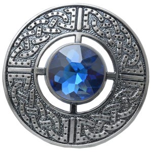 Puede incluir: Broche redondo plateado con una gema azul facetada en el centro. El broche presenta un intrincado dise&ntilde;o de nudos celtas alrededor de la gema y el borde exterior. El dise&ntilde;o es detallado y sim&eacute;trico, con un acabado pulido.