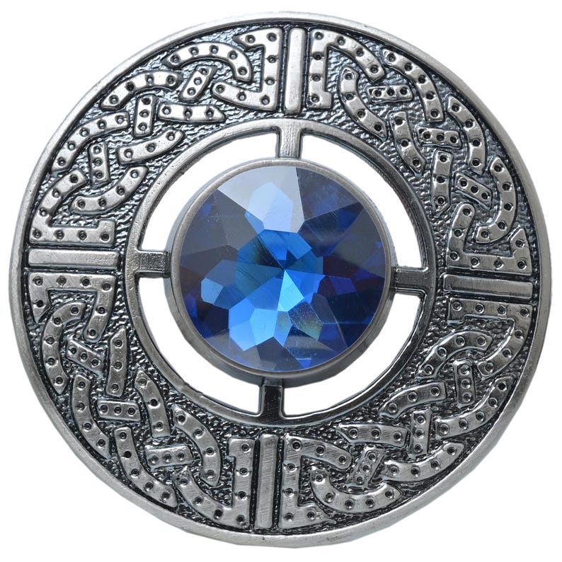 Puede incluir: Broche redondo plateado con una gema azul facetada en el centro. El broche presenta un intrincado dise&ntilde;o de nudos celtas alrededor de la gema y el borde exterior. El dise&ntilde;o es detallado y sim&eacute;trico, con un acabado pulido.