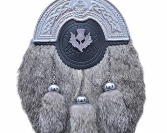 May include: A traditional Scottish sporran with a gray fur body. The sporran features a silver metal cantle with Celtic knotwork and a black leather flap with a thistle emblem. Three silver tassels hang from the bottom.