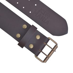 May include: A close-up of a brown leather belt with a brass buckle and rivets. The belt has the text "Kilt MANIA" embossed on the back. The belt is designed for kilts and other traditional Scottish attire.
