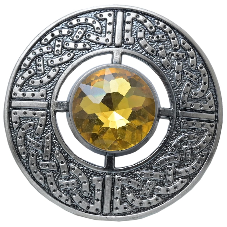 Puede incluir: Broche redondo plateado con un dise&ntilde;o de nudo celta en el borde exterior. El broche presenta una gran gema amarilla facetada en el centro. El dise&ntilde;o es sim&eacute;trico y detallado.