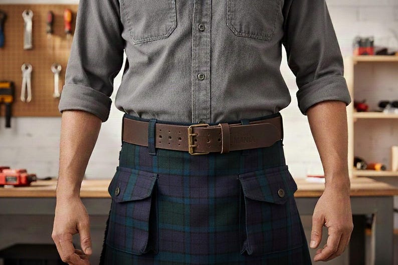 May include: A brown leather belt with a brass buckle is worn over a blue and green plaid kilt. The person is wearing a gray button-down shirt with rolled-up sleeves. The background includes a wooden workbench and tools.