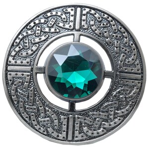 Puede incluir: Broche redondo plateado con un intrincado dise&ntilde;o de nudo celta. El broche presenta una gran gema verde esmeralda facetada en su centro. El dise&ntilde;o es detallado y sim&eacute;trico, con un acabado pulido.