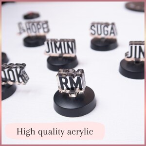May include: A collection of small, clear acrylic name plates featuring black text on white backgrounds, each mounted on a circular black base. The visible names include RM, J-HOPE, SUGA, JIMIN, JIN, and a partial view of COOK. The text High quality acrylic is present.